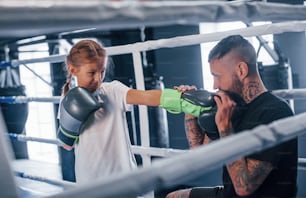 Ter sparring um com o outro no ringue de boxe. Jovem treinador de boxe tatuado ensina garotinha fofa na academia.