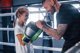 Having sparring with each other on the boxing ring. Young tattooed boxing coach teaches cute little girl in the gym.