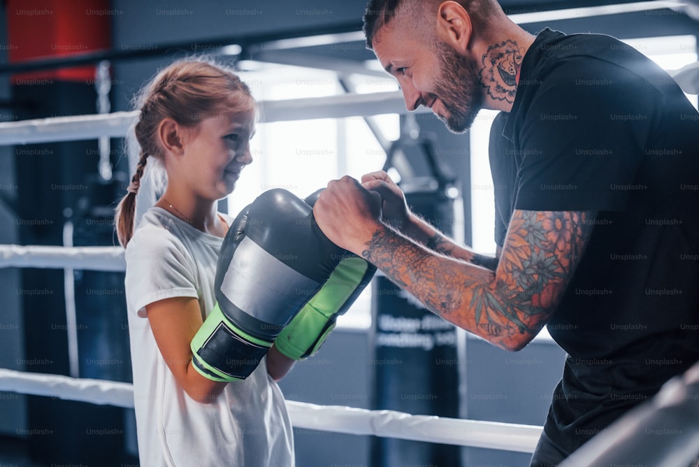 Having sparring with each other on the boxing ring. Young tattooed boxing coach teaches cute little girl in the gym.