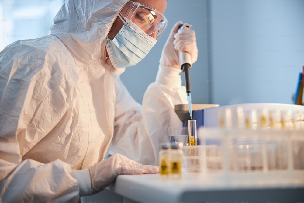 Side view of a serious lab technician in a hazmat suit immersing an automated pipette tip into the liquid