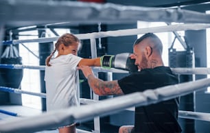 Having sparring with each other on the boxing ring. Young tattooed boxing coach teaches cute little girl in the gym.