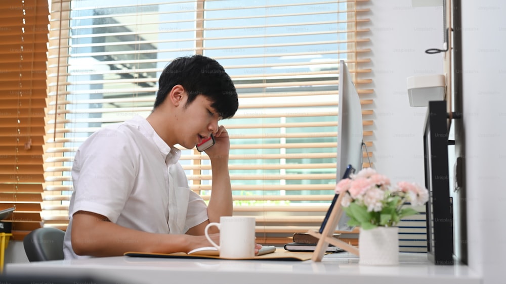 Side view of young asian man talking on mobile phone and working on computer at comfortable office.