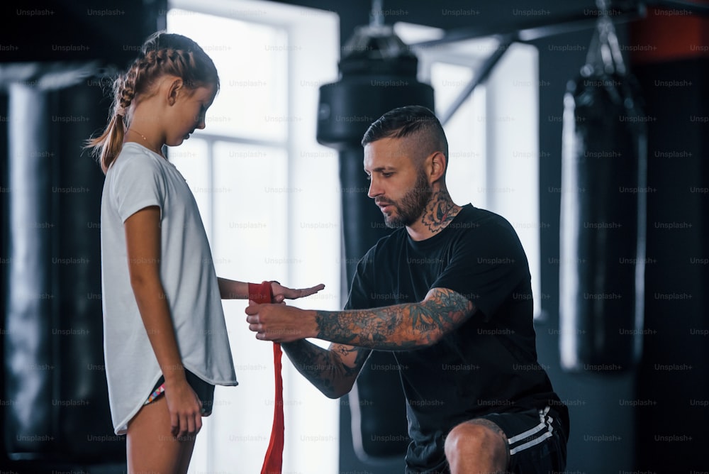 Helping to wear the bandages. Young tattooed boxing coach teaches cute little girl in the gym.