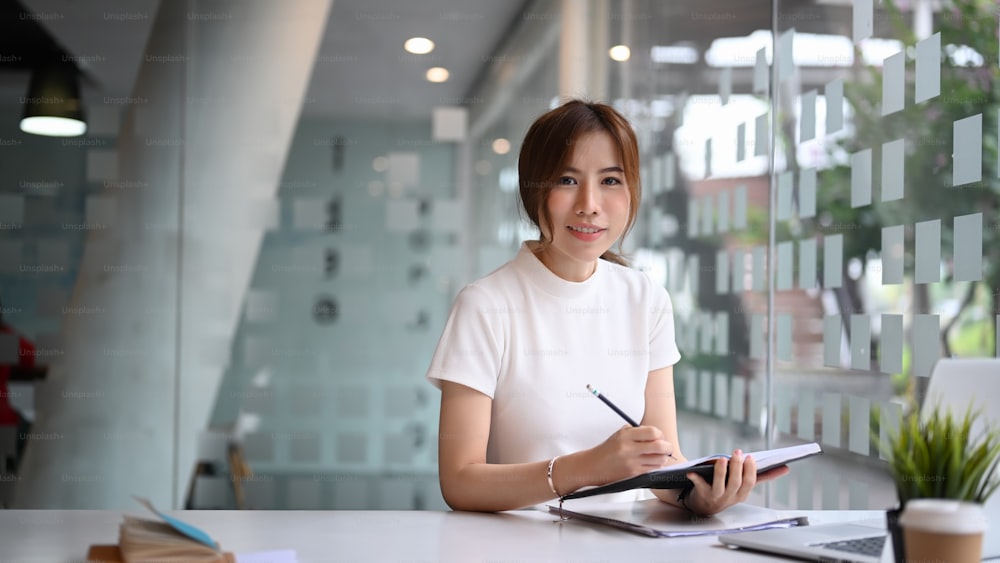 Mujer de negocios exitosa sosteniendo un cuaderno y sonriendo a la cámara mientras está sentada en un lugar de trabajo moderno.