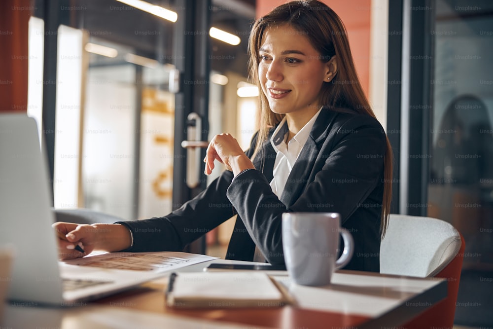 Focused pleased dark-haired lady with a ball-point pen in one hand staring at her laptop