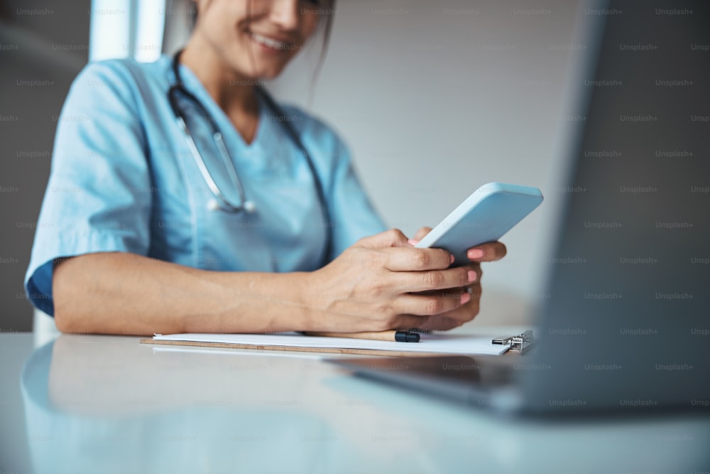 Close up of joyful young woman physician texting message on smartphone and smiling while sitting at the table with clipboard and laptop