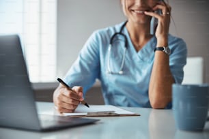 Close up of cheerful young woman physician having phone conversation and smiling while sitting at the table and taking notes