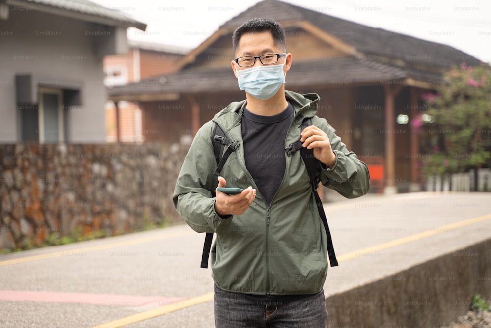 若いバックパッカーの男性は一人旅で、スマートフォンを使ってマスクと眼鏡を着用して方法を見つけています。