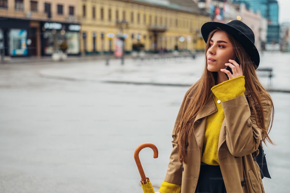 Mujer joven y hermosa que usa un teléfono inteligente y sostiene un paraguas amarillo mientras camina por la ciudad en un día lluvioso