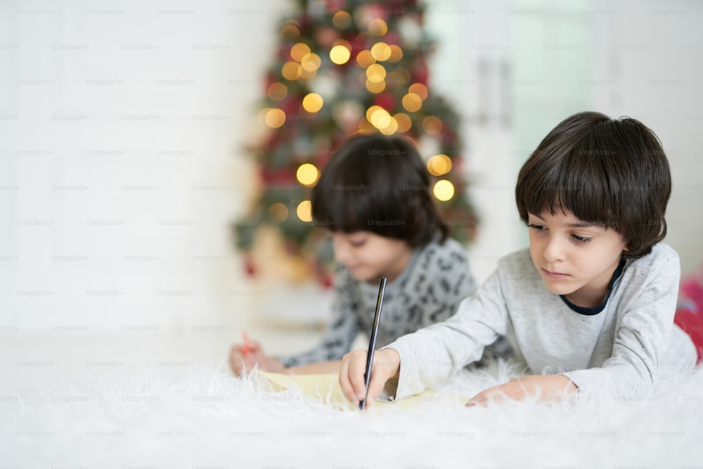 クリスマスの飾り付けをした家の床に寝そべりながら、カラフルな鉛筆で絵を描いている双子の2人の小さなラテンの男の子。子供時代、家族、冬休みのコンセプト