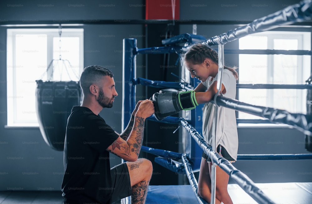 Conception of assistance. Young tattooed boxing coach teaches cute little girl in the gym.
