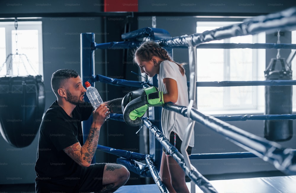 Concepção de assistência. Jovem treinador de boxe tatuado ensina garotinha fofa na academia.