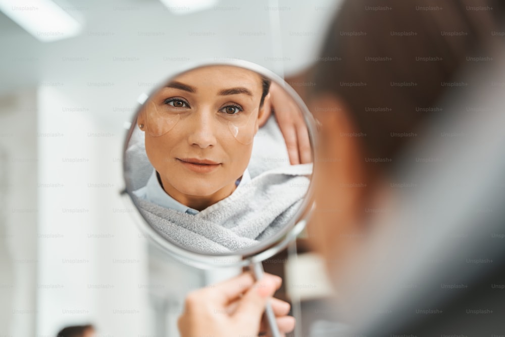 Ritratto di un cliente del salone di bellezza focalizzato con le bende sotto gli occhi in idrogel che esaminano il suo riflesso