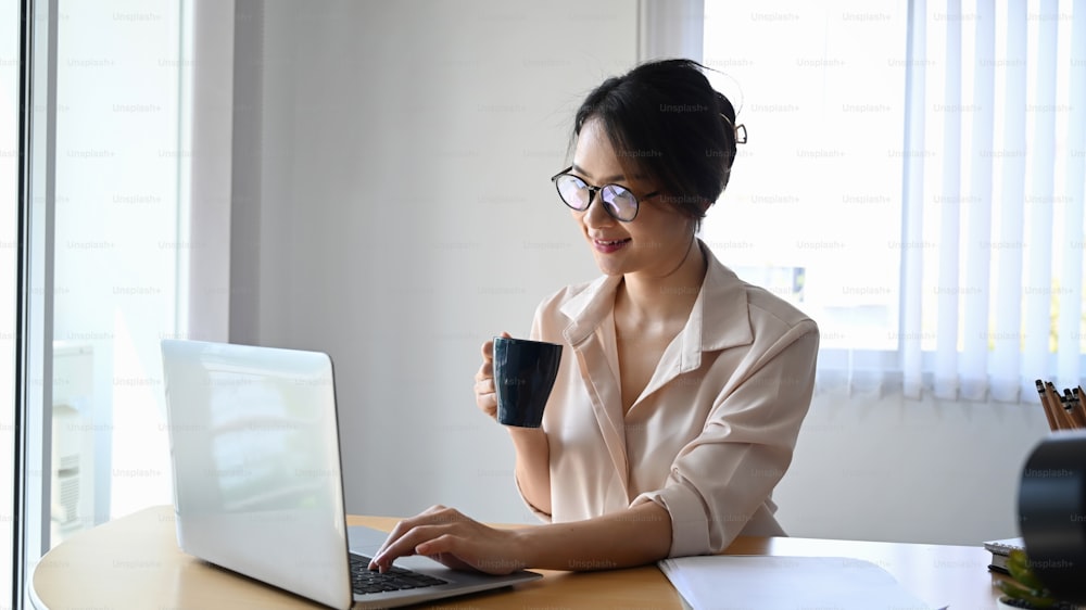 Smiling businesswoman in eyeglasses holding coffee cup and checking email on laptop computer.