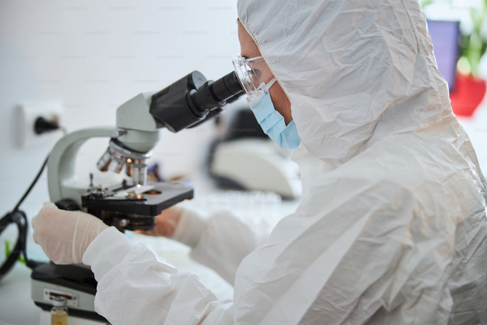 Side view of a woman in a face mask and safety goggles looking at cells under the microscope