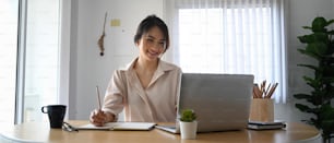 Smiling female employee making note on notebook and looking at screen of laptop computer.