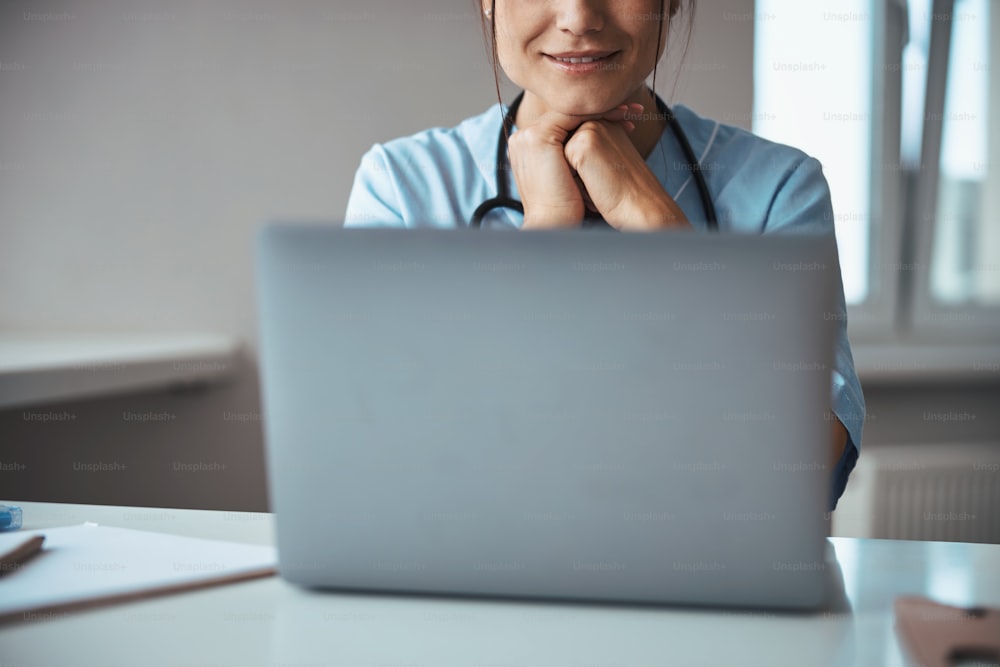 Close up of joyful young woman medical worker sitting at the table with modern notebook