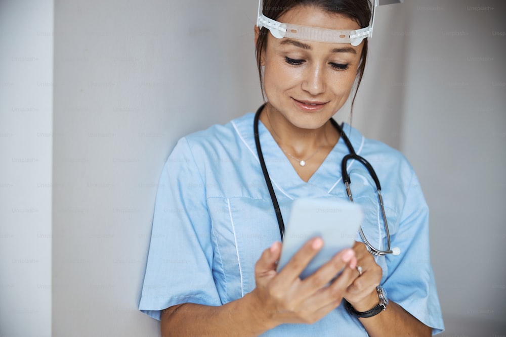 スマートフォンでメッセージをテキストメッセージで送信しながら、保護フェイスマスクと医療制服を着用している美しい若い女性医師
