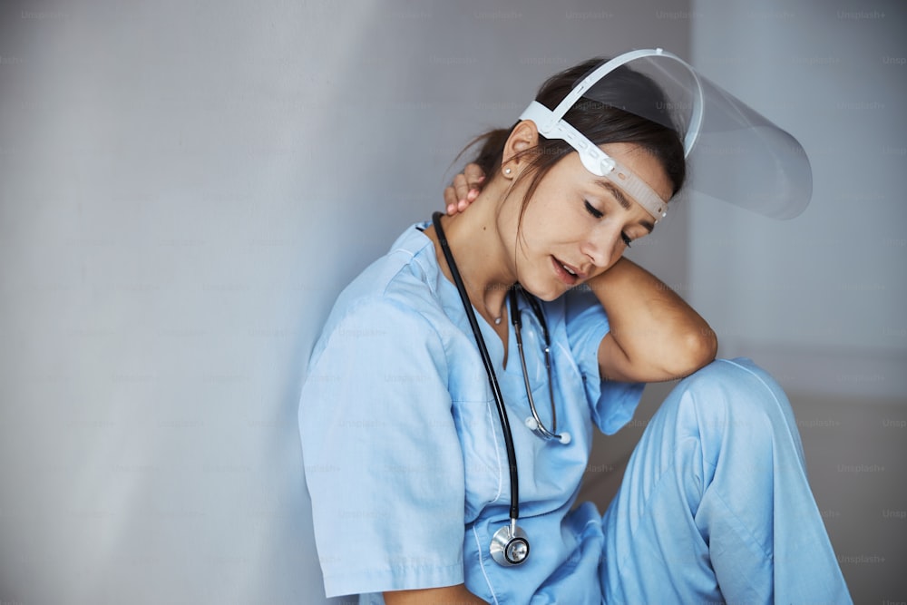 Jovem trabalhadora médica cansada usando máscara facial protetora e uniforme hospitalar enquanto sofre de fadiga