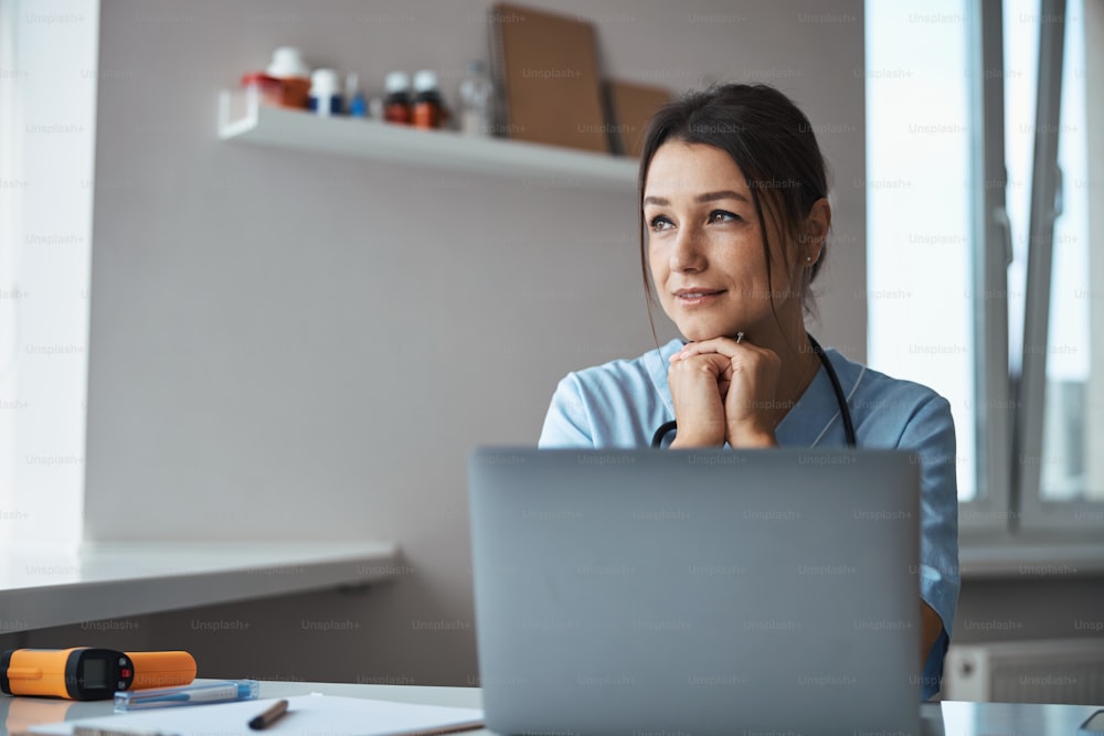 Charming young woman medical worker sitting at the table with modern notebook and looking out the window