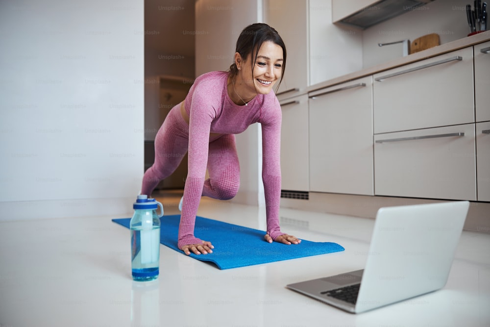 Beautiful sporty lady doing exercise and smiling while watching online training on notebook