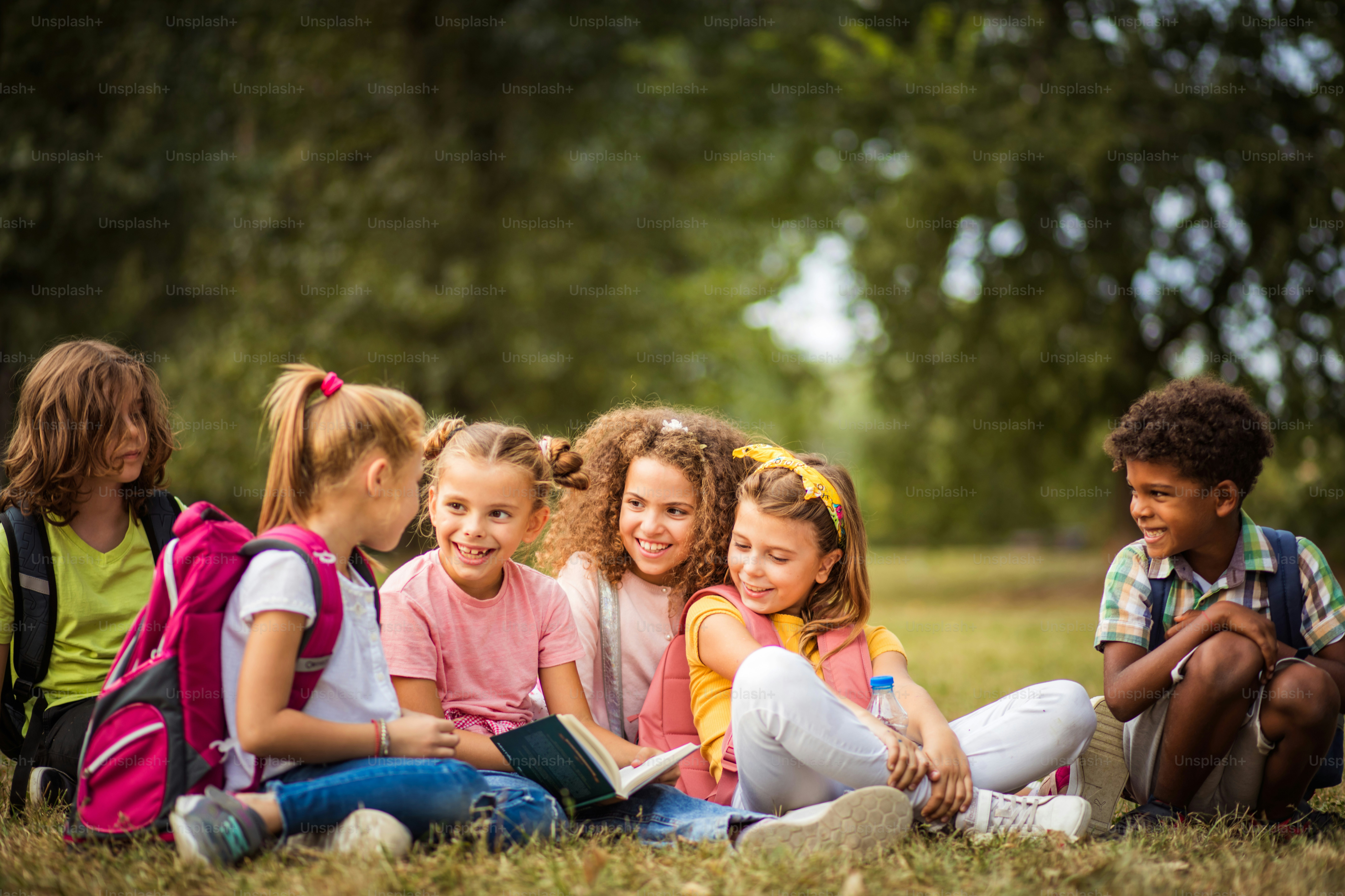 Große Gruppe von Schulkindern, die Spaß in der Natur haben. Kleines Mädchen liest ihren Freunden ein Buch vor.