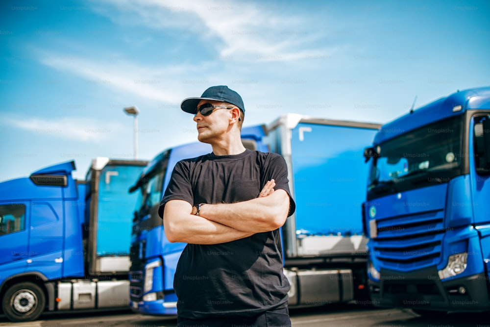 모자와 선글라스를 쓴 전문 트럭 운전사가 크고 현대적인 트럭 함대 앞에 자신 있게 서 있습니다. 밝고 화창한 날. 사람과 교통 개념.