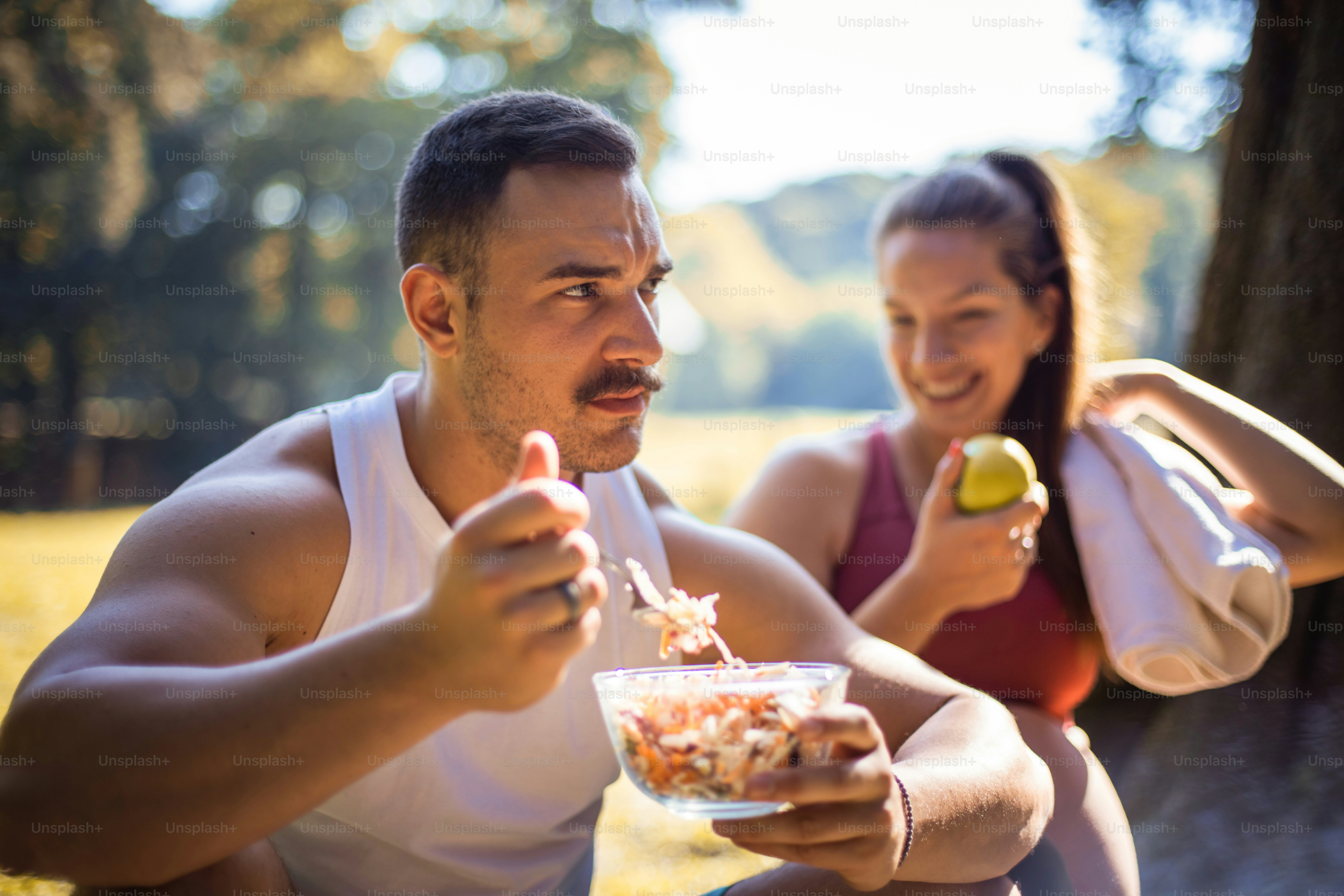 Couple in the park having break and eating healthy meal. Salad and apple. Focus is on man.