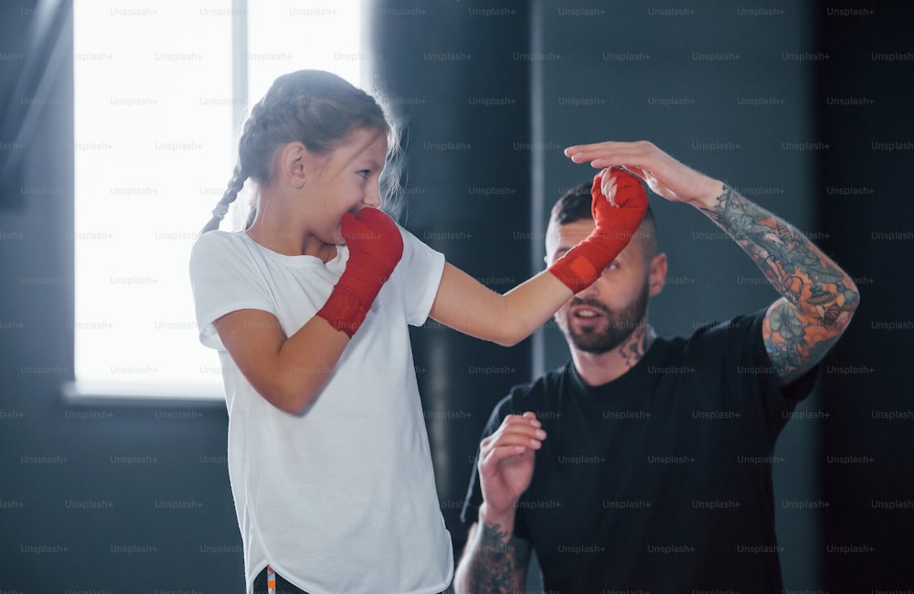 Basic punches. Young tattooed boxing coach teaches cute little girl in the gym.