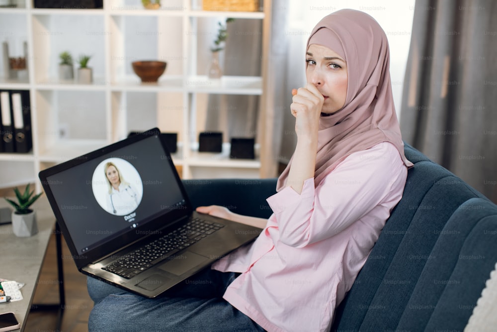 Sick Arabian woman in headscarf, having sore throat, pain and cough, sitting on couch at home and calling for consultation with medical practitioner, using laptop pc. Online doctor appointment.