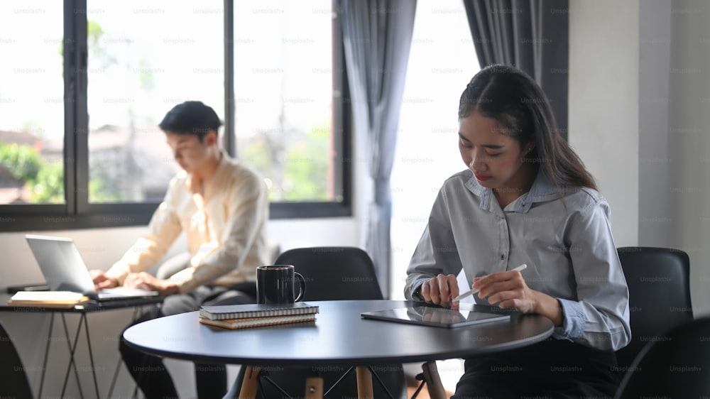 Junge asiatische Mitarbeiterin, die mit einem digitalen Tablet arbeitet, während sie mit ihrer Kollegin im Büroraum sitzt.