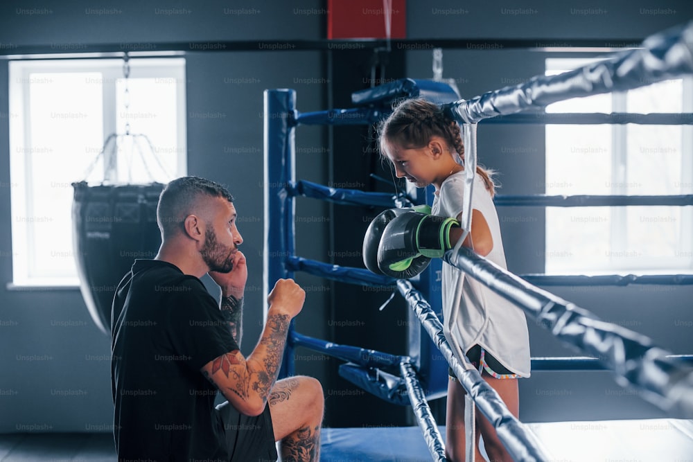 Konzeption von Hilfestellungen. Junger tätowierter Boxtrainer unterrichtet süßes kleines Mädchen im Fitnessstudio.