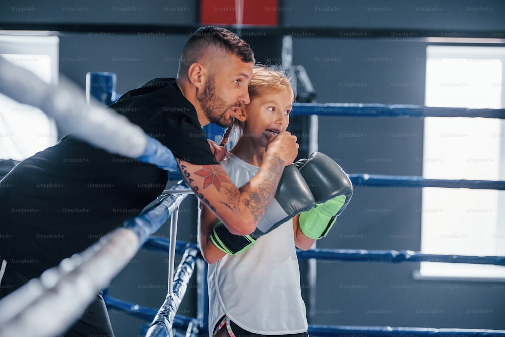 Standing on the boxing ring. Young tattooed boxing coach teaches cute little girl in the gym.