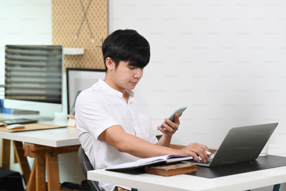 Young man employee working with laptop computer and using mobile phone in home office.