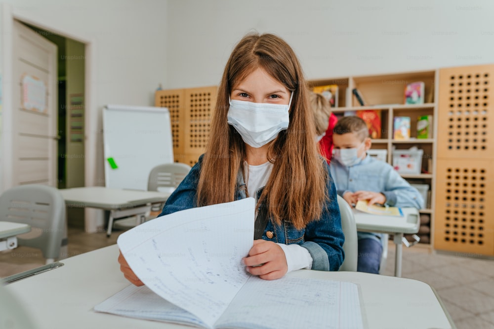 Grundschüler, die während der COVID-19-Pandemie im Klassenzimmer lernen. Mädchen mit Schutzmaske schaut in die Kamera. Selektiver Fokus
