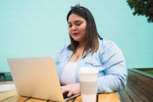 Portrait of young pluse size woman using her laptop while sitting outdoors at coffee shop. Technology and lifestyle concept.