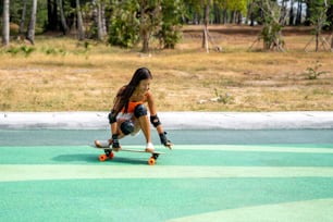 ビーチのそばのスケートボードパークでスケートをしている安全なスケートボードの膝パッドを持つ魅力的なアジアの女性。幸せな女性は、夏のアウトドアアクティブライフスタイルを楽しんで、公園でエクストリームスポーツサーフスケートをします。