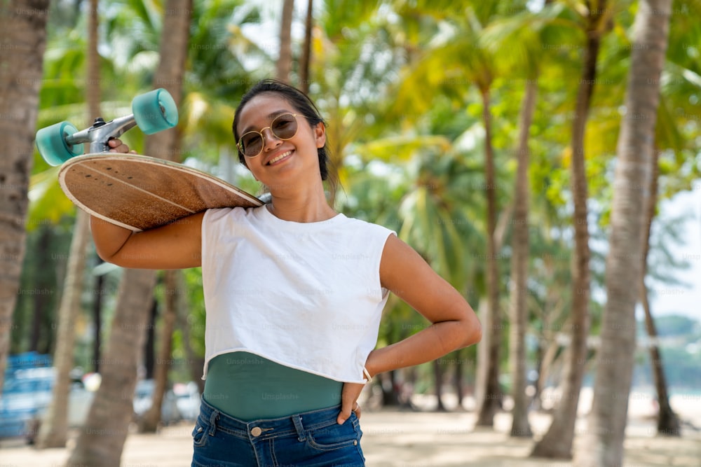 Retrato da mulher asiática sorridente atraente segurando skate andando na praia no dia ensolarado do verão. Confiança feminina desfrutar e ter divertido estilo de vida de atividade ao ar livre e esportes radicais surf skate