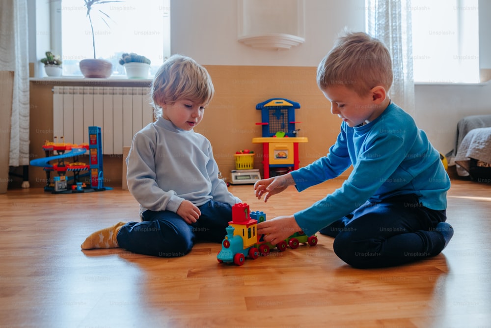 Two brothers 3 and 6 years boys playing train colorful blocks toy in the kids room sitting on the floor. Selective focus.
