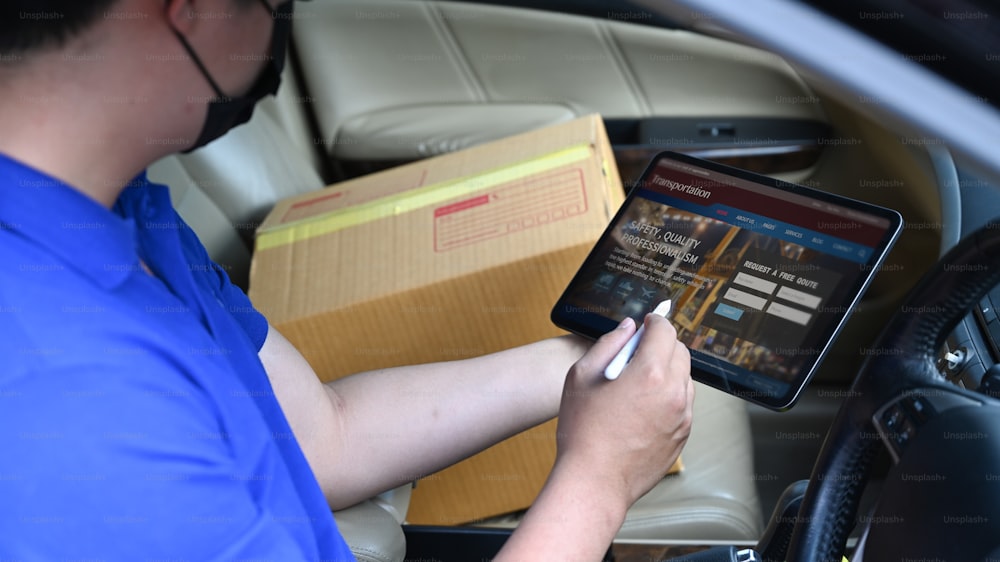 Autista del fattorino che utilizza la tavoletta digitale mentre è seduto nel furgone con la scatola di cartone. Servizio di consegna e concetto di spedizione.