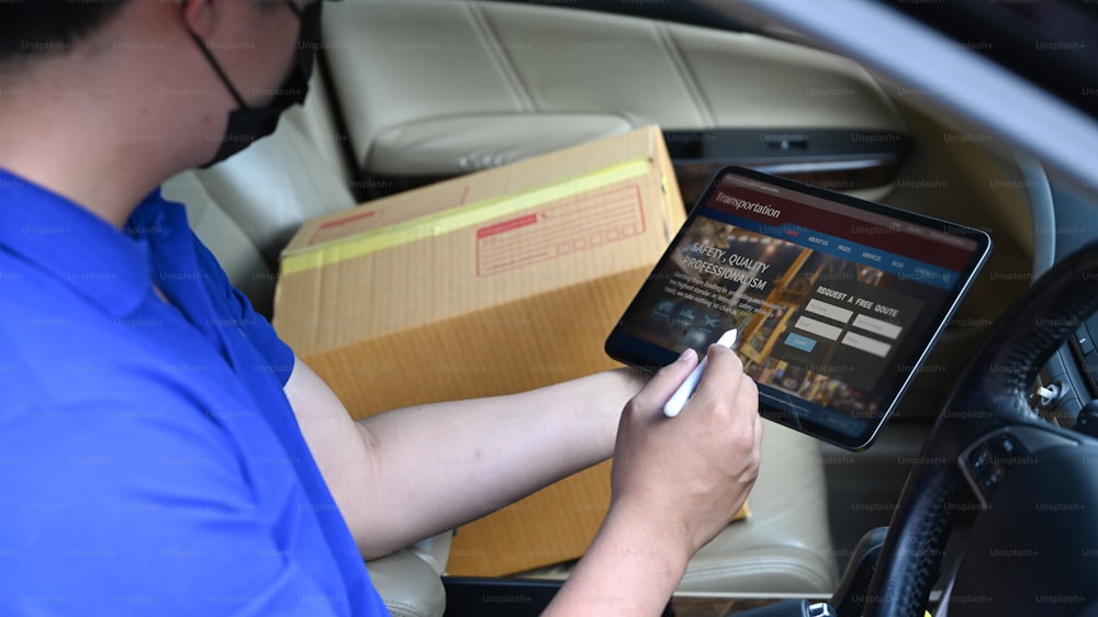 Delivery man driver using digital tablet while sitting in van with cardboard box. Delivery service and shipping concept.