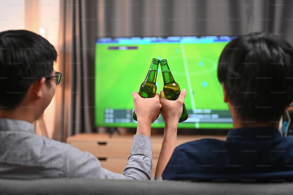 Rückansicht von zwei Männern, die ihre Gläser Bier anstoßen und auf dem Sofa Fußball anfeuern.