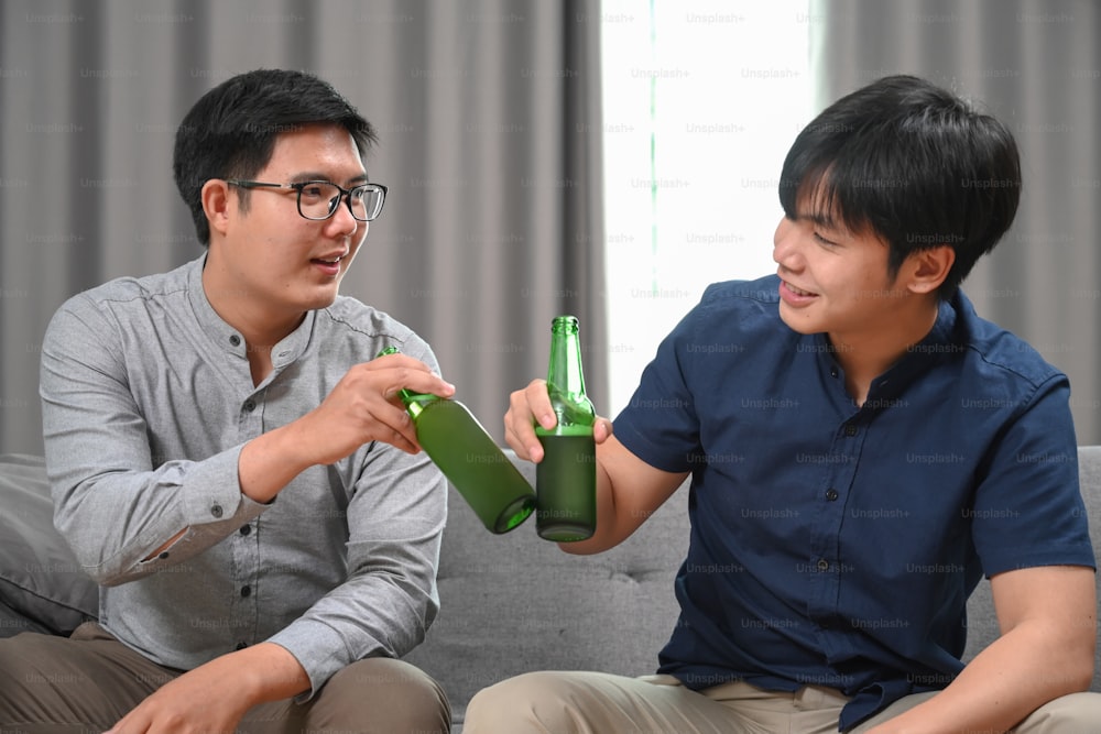 Due uomini asiatici che fanno tintinnare i loro bicchieri di birra mentre sono seduti sul divano di casa.