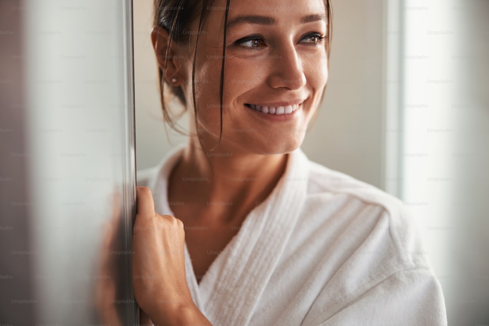 Cropped head portrait of happy smiling pretty business woman in white bathrobe looking away while standing in room indoors