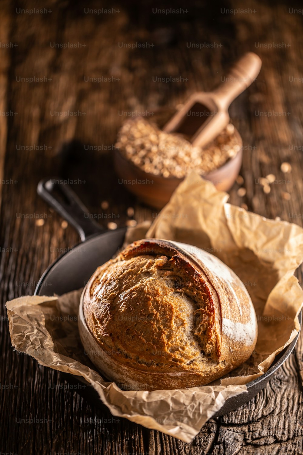 Freshly baked crunchy rustic yeast bread lying on the baking paper in the alloy pan wooden bowl full of wheat grains wooden scoop stitched deeply into it.