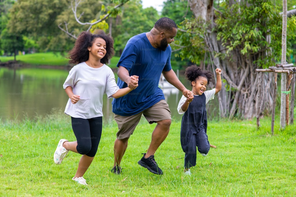 Happy mixed race family in park. African father carrying and holding hand two little daughter walking together in garden. Dad and cute child girl kid enjoy and having fun outdoor lifestyle activity