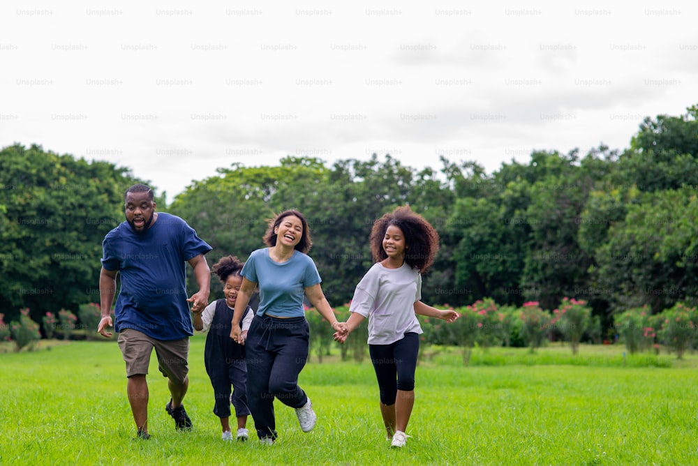 Happy mixed race family in park. African father and Asian mother with two little daughter holding hands walking together in garden. Mom and dad with child girl kid enjoy and having fun in outdoor lifestyle activity.