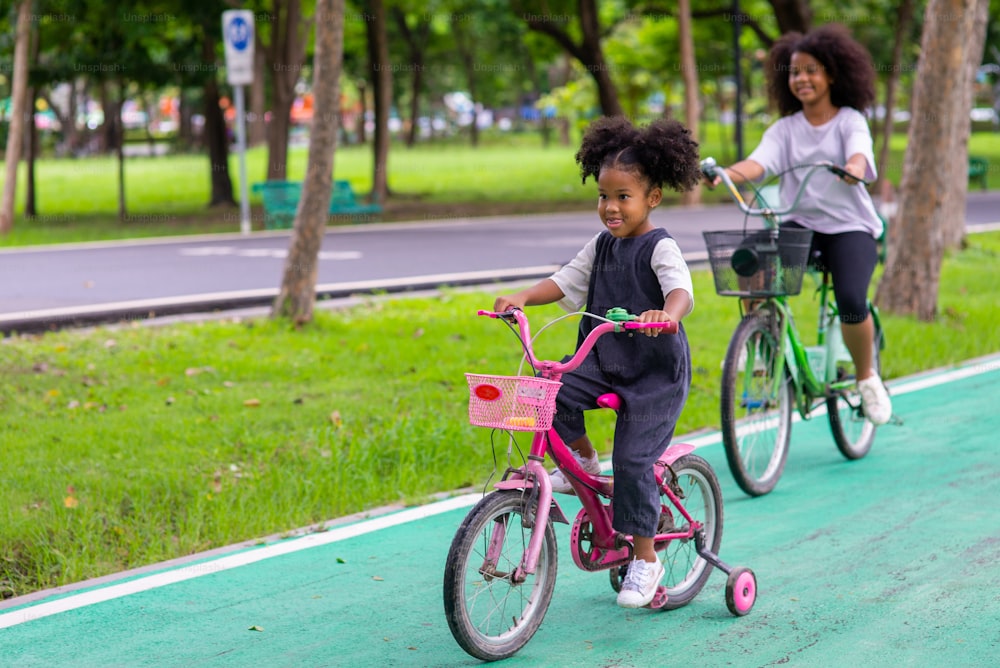 Happy mixed race family in park. Two Adorable little sibling sister learning ride bicycle together in public park. Adorable child girl kid enjoy and having fun outdoor lifestyle activity vacation.