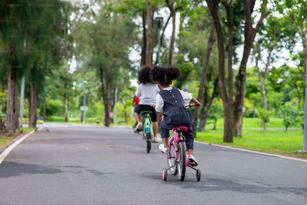 Happy mixed race family in park. Two Adorable little sibling sister learning ride bicycle together in public park. Adorable child girl kid enjoy and having fun outdoor lifestyle activity vacation.