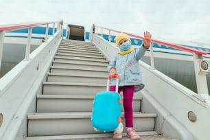 Little girl wearing protective face mask holding suitcase waving hand near the plane. Travel and tourism after the end of lockdown. New normal after Pandemic COVID-19 concept. Selective focus.